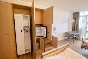 Patientzimmer mit Esstisch und Essstühle. Offene Kommode mit integrierten Kühlschrank, Mülleimer und fahrbahren Patientenschrank.