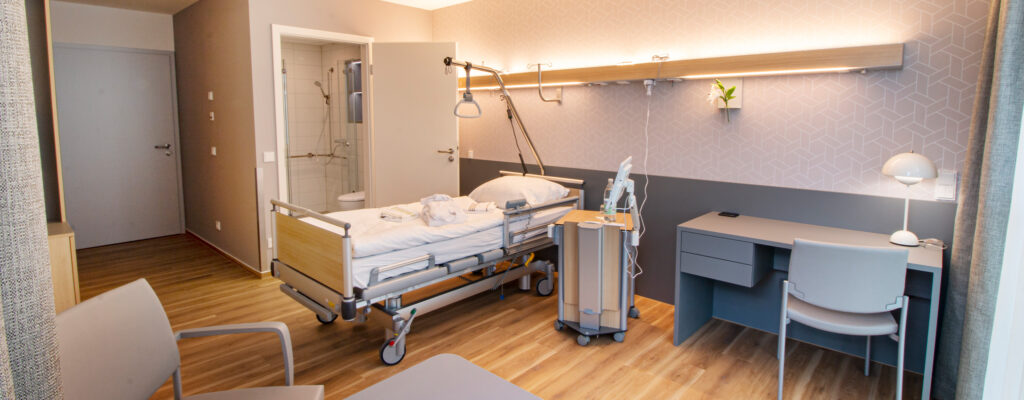 Patientenzimmer mit Patientenbett, Badezimmer, Sitzecke mit Tisch und Stuhl, Schreibtisch mit Stuhl, fahrbarer Nachttisch